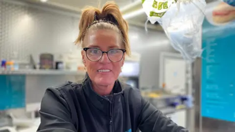 A woman with glasses and hair tied back stands inside a snack van, smiling at the camera